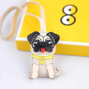 May include: A beige leather pug dog keychain with black ears, nose, and a red tongue. The pug has a yellow collar and a small yellow tag. The keychain is attached to a beige strap. The background is yellow.