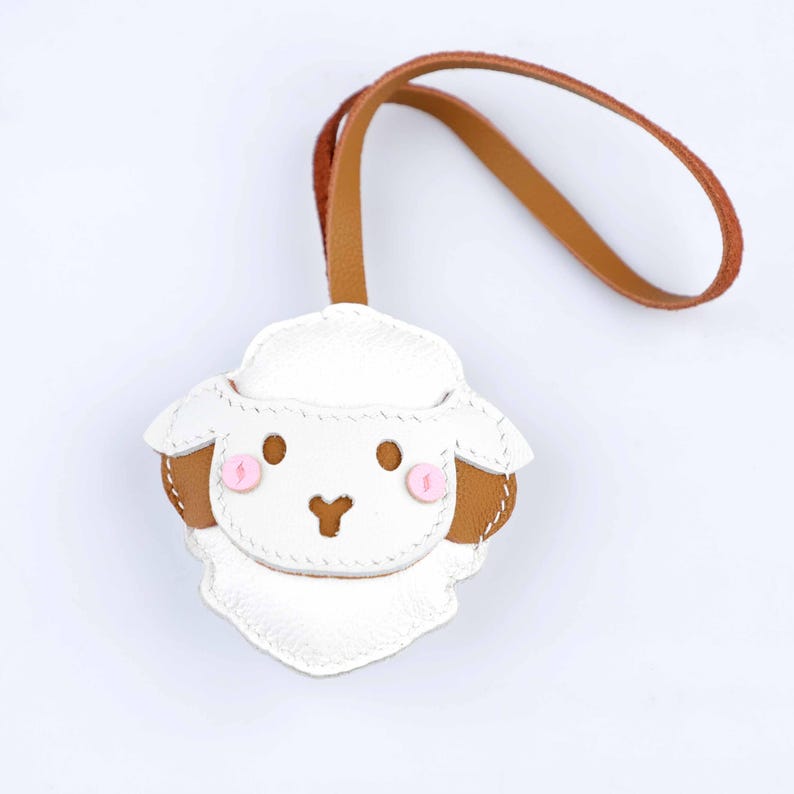 May include: A white and brown leather sheep keychain with a brown leather loop. The sheep has pink cheeks and a stitched design. The keychain is a cute accessory.