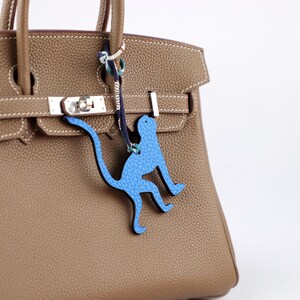 Puede incluir: Un bolso de mano de cuero marrón claro con un cierre plateado y un charm de cuero azul con forma de mono colgando del asa. El charm está sujeto con un cordón azul marino y blanco.