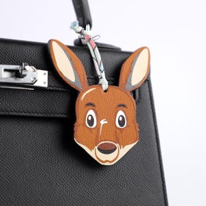 May include: A brown leather bunny charm with a cartoon-like face, attached to a black handbag. The bunny has large ears, a white muzzle, and big eyes. A patterned fabric cord connects the charm to the bag's handle.