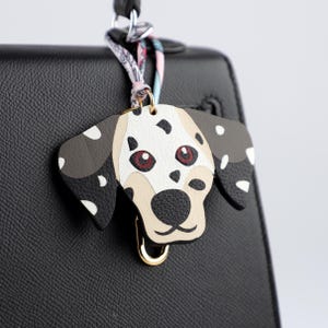 May include: A Dalmatian dog-shaped bag charm with a gold-colored clasp. The charm features a white base with black spots, tan muzzle, and red eyes. It is attached to a black handbag with a patterned scarf.