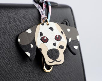 May include: A Dalmatian dog-shaped bag charm with a gold-colored clasp. The charm features a white base with black spots, tan muzzle, and red eyes. It is attached to a black handbag with a patterned scarf.