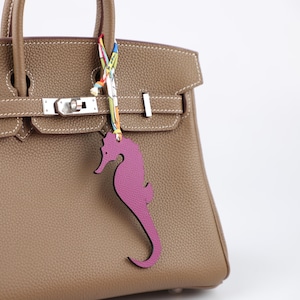 May include: A taupe leather handbag with silver hardware and a decorative seahorse charm. The charm is a vibrant magenta color and hangs from a colorful patterned ribbon. The bag is a classic style.