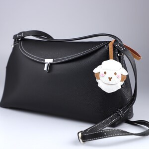 May include: Black leather crossbody bag with white stitching and a silver clasp. The bag features a detachable strap and a sheep-shaped charm with brown and pink accents.