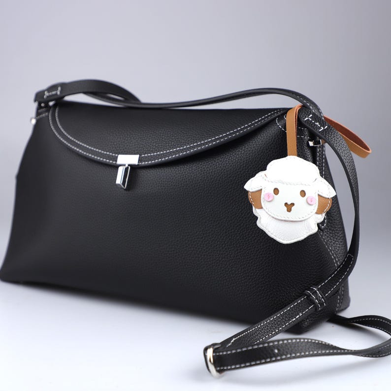 May include: Black leather crossbody bag with white stitching and a silver clasp. The bag features a detachable strap and a sheep-shaped charm with brown and pink accents.