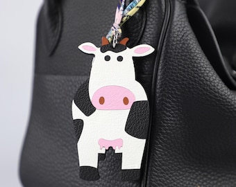 May include: A cow-shaped leather bag charm with a white body, black spots, and a pink face and udder. The charm is attached to a black leather bag with a colorful patterned ribbon.
