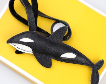 May include: A black and white leather orca keychain with a black strap. The orca has white patches and is stitched with white thread. The background is a bright yellow.