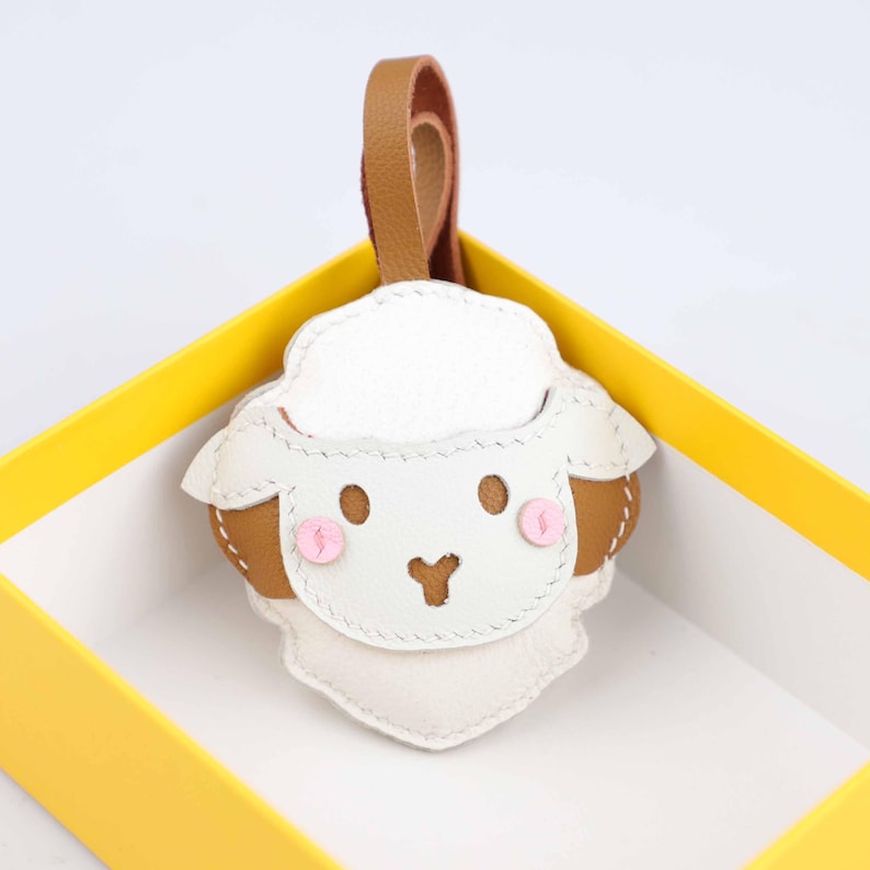 May include: A white and brown leather sheep keychain with a brown leather loop. The sheep has pink cheeks and a stitched design. The keychain is inside a yellow box.