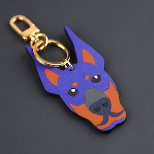 May include: A leather keychain in the shape of a Doberman dog's head. The dog is depicted in blue with orange markings. It has a gold-coloured clasp and ring for attaching to keys or bags.