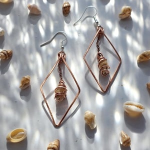 Op de afbeelding: Een paar oorbellen van koperkleurig draad met een ruitvormig frame. Elke oorbel heeft een kleine, beige spiraalvormige schelp die aan een kort kettinkje in het midden van de geometrische draadstructuur hangt, bevestigd aan een zilverkleurige haak.