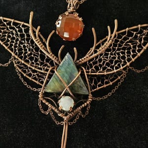Op de afbeelding: Een ketting van koperdraad met een driehoekige labradoriet, een ronde oranje gefacetteerde edelsteen en een kleine witte cabochon. Het ontwerp bevat ingewikkelde vleugels van draadgaas, delicate kettingaccenten en een koperen ketting.