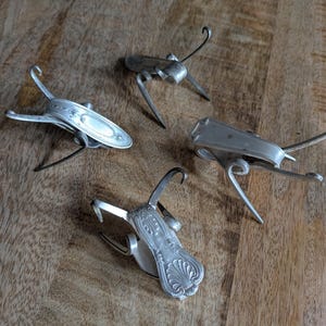 May include: Four handcrafted metal insect sculptures made from repurposed silverware. Each insect features a unique design, with curved legs and antennae. The silverware insects are arranged on a wooden surface, showcasing their intricate details and creative reuse.