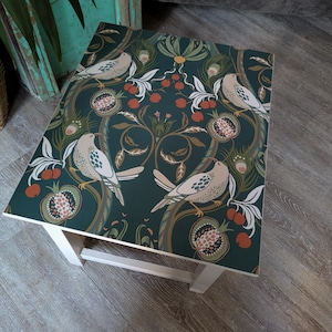 May include: A square side table featuring a dark green surface with a repeating pattern of light-coloured birds, pomegranates, and peacock feathers. The table has a simple white frame and four legs, providing a flat surface for display.