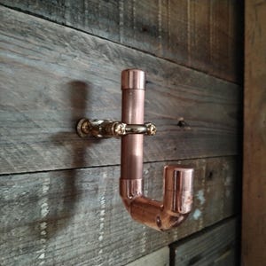 Puede incluir: Un gancho en forma de J de color cobre con herrajes de latón, montado sobre una superficie de madera envejecida. El gancho está construido con tuberías y accesorios de cobre, mostrando un diseño industrial. Los herrajes de latón añaden contraste.