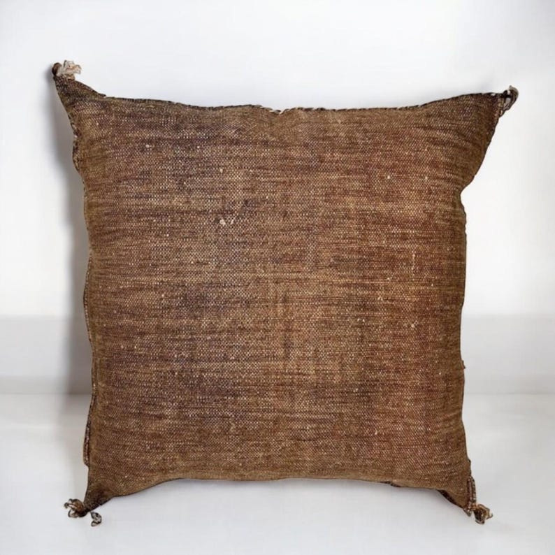 Puede incluir: Un coj&iacute;n decorativo cuadrado, marr&oacute;n, con un aspecto texturizado y tejido. El coj&iacute;n tiene esquinas puntiagudas y peque&ntilde;as borlas en cada esquina. La tela tiene un aspecto ligeramente desgastado, con variaciones en el color marr&oacute;n.