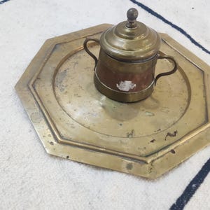 May include: An octagonal brass tray with a weathered patina and a central two-handled lidded container. The container features a polished brass lid with a knob handle, a copper-toned body, and a small white adhesive label on the front surface.
