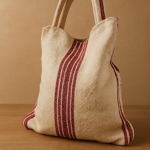 May include: A beige tote bag with red stripes. The bag has two handles and is made of a woven fabric.