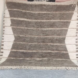May include: A large, rectangular rug with a geometric design. The rug features alternating bands of brown and white, with a diamond-shaped pattern in the center. The rug has a fringed edge and is made of a textured material.