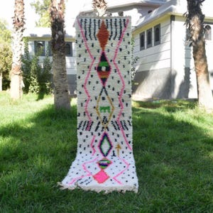 Op de afbeelding: Een rechthoekig, gebroken wit tapijt met een geometrisch patroon. Het ontwerp bevat zwarte, roze, oranje, groene en gele ruitvormen en accenten. Het tapijt wordt buiten op groen gras getoond.