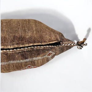 Puede incluir: Un coj&iacute;n rectangular marr&oacute;n con una textura tejida. Cuenta con una cremallera negra y patrones geom&eacute;tricos decorativos blancos y rojos en el borde. Una peque&ntilde;a borla est&aacute; unida al tirador de la cremallera.