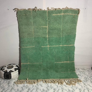 Könnte beinhalten: Ein großer, rechteckiger grüner Teppich mit strukturierter Oberfläche und geometrischem Design. Der Teppich hat hellbeige Linien und Fransen an den Rändern. Ein dekorativer Pouf mit schwarz-weißen Mustern ist unten links zu sehen.