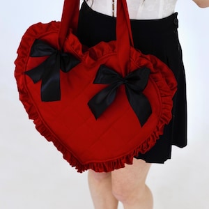 May include: A red heart-shaped handbag with black bows and frilled edges. The bag has a quilted pattern and two handles. The bag is held by a person wearing a white top and black skirt.