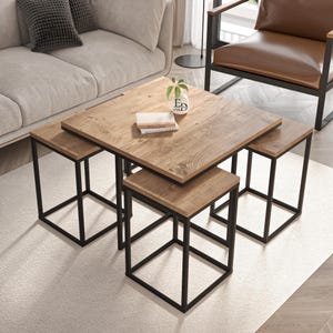 Op de afbeelding: Een vierkante houten salontafel met een natuurlijke houtnerfafwerking en een zwart metalen frame, vergezeld van twee bijpassende vierkante krukjes die eronder kunnen worden geschoven. Een kleine witte vaas met een plant en een boek staan op het tafelblad.