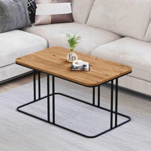 Peut inclure: Une table basse rectangulaire avec un plateau en bois clair au grain apparent et une structure en métal noir. Le cadre se compose de fins supports verticaux reliés à une base rectangulaire continue. Un petit vase blanc et un livre sont posés dessus.
