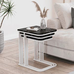 Peut inclure: Un ensemble de trois tables gigognes avec des plateaux carrés à motif marbre noir et des structures en métal blanc. Les tables sont empilées, chacune ayant une base en forme de C. Un petit vase avec les lettres ED est sur le dessus.