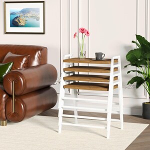 Puede incluir: Un juego de cuatro mesas nido con superficies rectangulares de madera y estructura de metal blanco. Las mesas están apiladas verticalmente, con un florero de vidrio transparente con flores rosas y una taza gris sobre la superficie.