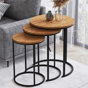 Peut inclure: Un ensemble de trois tables gigognes avec des plateaux ronds en bois clair à grain apparent et des structures en métal noir. Chaque table possède une base circulaire et trois pieds verticaux. Un vase en verre avec des tiges séchées est posé.