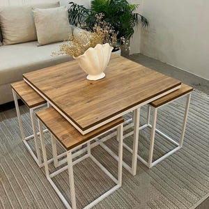 Op de afbeelding: Een set van vier nesttafels met vierkante, houtnerf-achtige bladen en witte metalen frames. De grootste tafel is ongeveer 76 cm vierkant. Een witte, schelpvormige vaas bevat gedroogde bloemen.