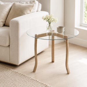 Peut inclure: Une table d'appoint ronde dotée d'un plateau en verre transparent reposant sur trois pieds en bois clair incurvés. La table supporte un petit vase en verre contenant des fleurs blanches et une tasse en céramique beige au design épuré.