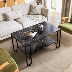 Peut inclure: Une table basse rectangulaire avec un plateau en faux marbre noir et une étagère inférieure assortie, soutenue par un cadre en métal noir. La surface présente des veines blanches et porte un vase blanc avec les lettres EB, un bol et des livres.