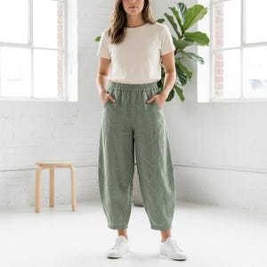 May include: A woman wearing a cream-colored t-shirt and olive green linen pants. The pants have an elastic waistband and a relaxed fit. She is also wearing white sneakers. The background includes a large plant and a white brick wall.
