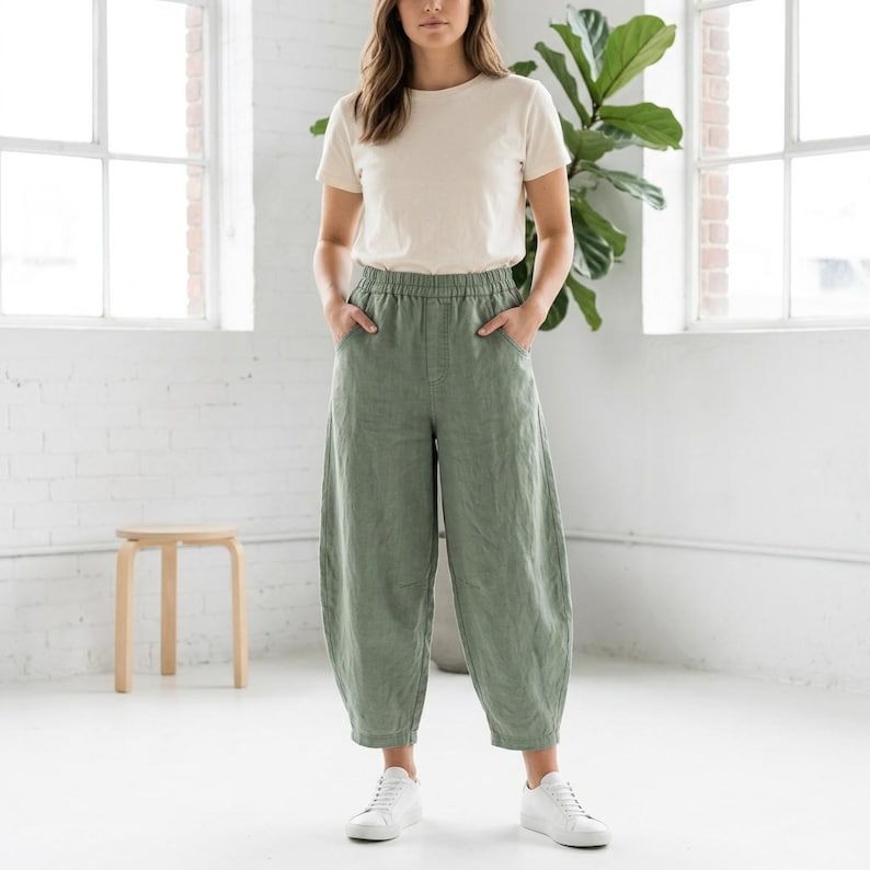 May include: A woman wearing a cream-colored t-shirt and olive green linen pants. The pants have an elastic waistband and a relaxed fit. She is also wearing white sneakers. The background includes a large plant and a white brick wall.