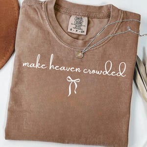 May include: A brown Comfort Colors t-shirt with the phrase "make heaven crowded" in white script. A silver necklace with a small pendant rests on the shirt. The shirt has a small tag at the top.
