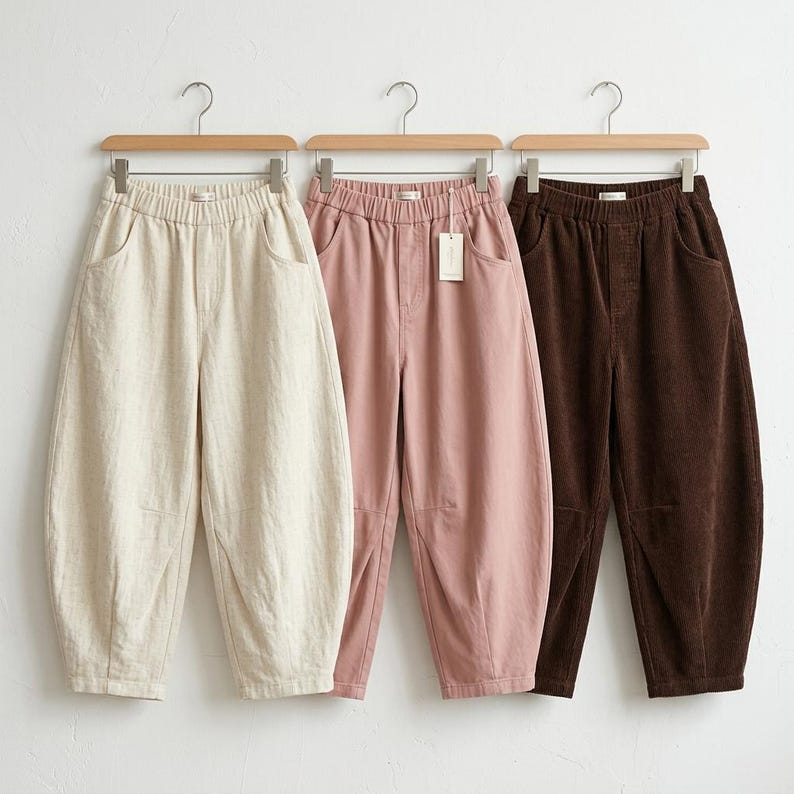 May include: Three pairs of casual pants in different colors hanging on wooden hangers. The pants are beige, pink, and brown. Each pair has an elastic waistband and pockets. The pants are made of a soft, comfortable material.