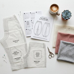 May include: An overhead shot of sewing supplies, including pattern pieces, fabric swatches, and a pair of scissors. The pattern is for "THE ARCADI BALLOON-LEG PANTS." A cup of coffee and a small succulent plant are also present, suggesting a crafting project.