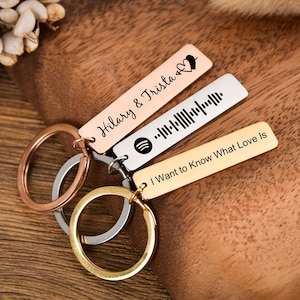 May include: A collection of personalized keychains in rose gold, silver, and gold tones. Each rectangular tag features custom text or a Spotify code. One keychain reads "Hilary & Trista" with a heart, another displays a Spotify code, and the third says "I Want to Know What Love Is."