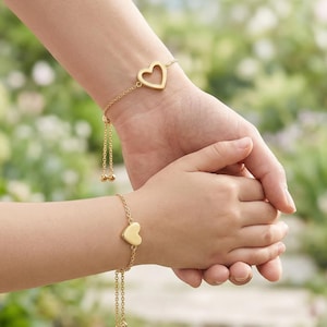 May include: Two gold-colored heart bracelets, one on an adult's wrist and the other on a child's wrist. The adult's bracelet features a heart-shaped cutout, while the child's has a solid heart. Both bracelets have delicate chain details.
