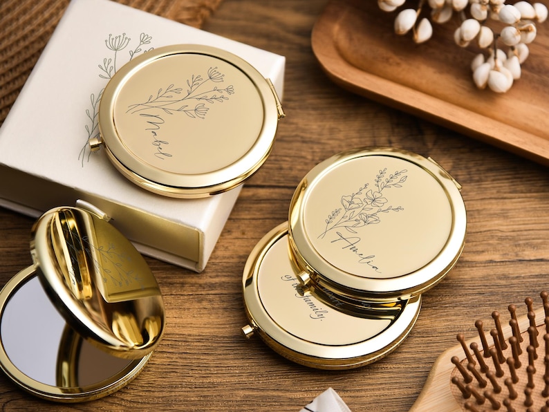 May include: Gold-colored compact mirrors with floral designs and personalized names. One mirror is open, revealing a reflective surface. The mirrors are on a wooden surface, with a white box and a wooden tray in the background.