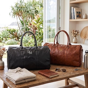Könnte beinhalten: Zwei große Reisetaschen, eine schwarze und eine braune, mit zwei Tragegriffen und personalisierten Monogrammbuchstaben. Die schwarze Tasche zeigt die Buchstaben DM, während die braune Tasche die Buchstaben CS aufweist. Beide Taschen haben eine rechteckige Form.