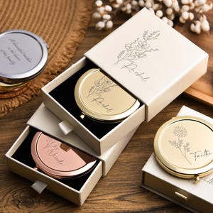 May include: A collection of personalized compact mirrors in gold, silver, and rose gold, some in open boxes. The mirrors have floral designs and names engraved on them. One mirror has the text "A beautiful mother is the heart of the family."