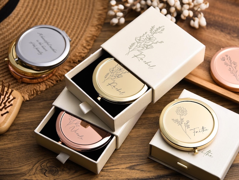 May include: A collection of personalized compact mirrors in gold, silver, and rose gold, some in open boxes. The mirrors have floral designs and names engraved on them. One mirror has the text "A beautiful mother is the heart of the family."
