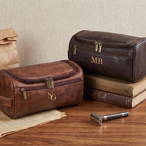 May include: Two rectangular leather toiletry bags with zippered closures and brass-toned hardware. One bag is light brown with the initials NG, and the other is dark brown with the initials MR. A metal safety razor is placed in the foreground.