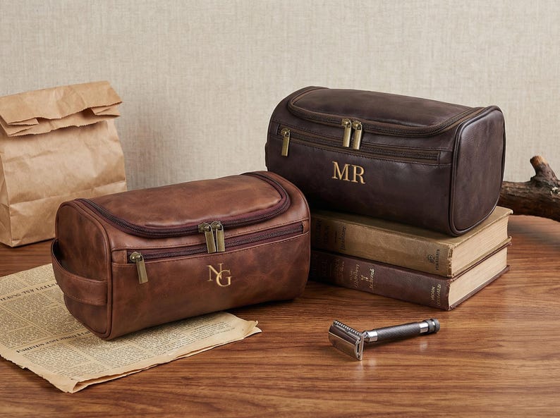 May include: Two rectangular leather toiletry bags with zippered closures and brass-toned hardware. One bag is light brown with the initials NG, and the other is dark brown with the initials MR. A metal safety razor is placed in the foreground.
