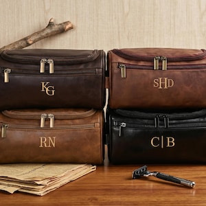 以下が含まれることがあります： 茶色と黒の色合いの革製化粧ポーチ4つ。それぞれに金色のジッパーとパーソナライズされたイニシャルが付いています。木製の表面にはカミソリが置かれています。バッグは積み重ねられており、さまざまな色とデザインが示されています。