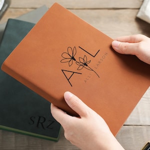 May include: A tan leather journal featuring the initials "AL" and a floral motif. The name "Alli Larson" is printed below the design. The journal is held open, revealing the pages. Other journals are visible in the background.
