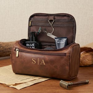 May include: A brown leather toiletry bag with a zipped opening and the initials S|A embossed on the front. The bag is open, revealing an interior with a metal hanging hook, a mesh pocket, and various personal care items inside.